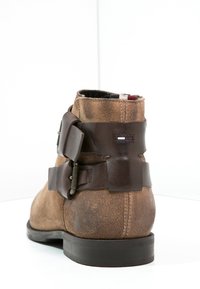 Stivaletto in suede marrone con punta tondeggiante, cinturini in pelle marrone con fibbia e suola in gomma nera. Presenta una piccola etichetta con logo.