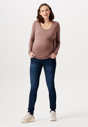Bluzka ciążowa z miękkiego, mauve'owego materiału z krojem typu wrap, połączona z ciemnoniebieskimi rurkami i jasnobeżowymi trampkami.