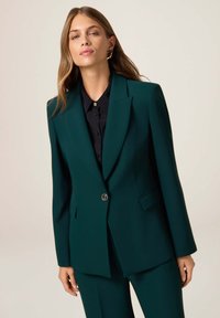 Blazer ajustado en azul verdoso con hombros estructurados, cierre de un solo botón y dos bolsillos frontales. Combinado con una camisa negra de botones.
