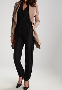 Nainen, jolla on päällään musta kietaisujumpsuit, jonka vyötärö on sidottu, beige vesiputous-kaarroke ja mustat kärkitossut, seisoo tasaista taustaa vasten.