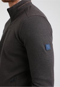 Giacca zip-up marrone scuro in tessuto a rete simile a un alveare, con un'etichetta blu sul maniche sinistro e un sottile tocco di colore al colletto.
