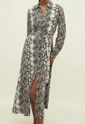 Vrouw draagt een midi-jurk met lange mouwen en ceintuur, met een slangenhuidprint in zwart, wit en beige, en een voorste split die één been laat zien.