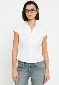 Λευκό πουκάμισο με κουμπιά στο μπροστινό μέρος και κοντά μανίκια, με κάθετες ρίγες στο πάνω μέρος και εφαρμοστή μέση, συμπληρωμένο με κλασικό γιακά.