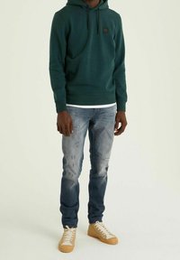 Felpa verde con cappuccio e logo nero, abbinata a jeans azzurri chiari strappati e sneakers beige. L'outfit presenta un fit casual.