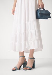 Ljusblå sandaletter med klack och ankelremmar, med en överdel i texturerat tyg och en klumpig klack, kombinerade med en vit klänning med lager på lager.