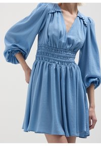 Robe bleu clair en tissu texturé, avec un col en V, une taille smockée et des manches bouffantes, ainsi qu'une jupe évasée tombant à mi-cuisse.