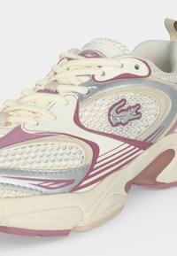 Baskets athlétiques crème, rose et argent avec des empiècements en mesh texturé, lacets à l'avant et logo crocodile sur le côté.