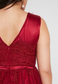 Dieprood jurk met een V-rugontwerp, ingewikkelde bloemenkantdetails op het bovenlichaam en een gladde satijnen textuur.