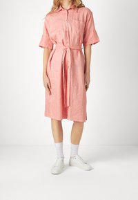 Abito camicia di lino rosa con vita elasticizzata, maniche corte e chiusura a bottoni. Abbinato a calzini bianchi e scarpe da ginnastica.