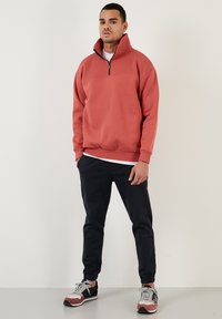 Rode sweatshirt met een halflange ritskraag, geribde manchetten en zoom. Draagt het met zwarte broek en multicolore sneakers, staande tegen een effen achtergrond.