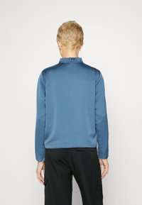 Top blu, a maniche lunghe con colletto arricciato e texture liscia, presenta una vestibilità comoda e una leggera lucentezza, abbinato a pantaloni cargo scuri.