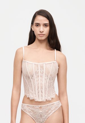 Une femme porte un haut corset en dentelle rose pâle et des sous-vêtements assortis, sur un fond clair et uni.