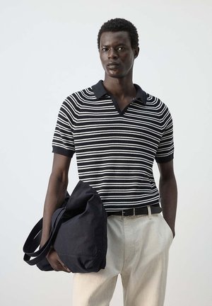 Homme portant un polo rayé noir et blanc et un pantalon clair, tenant un grand sac noir avec une main dans la poche.