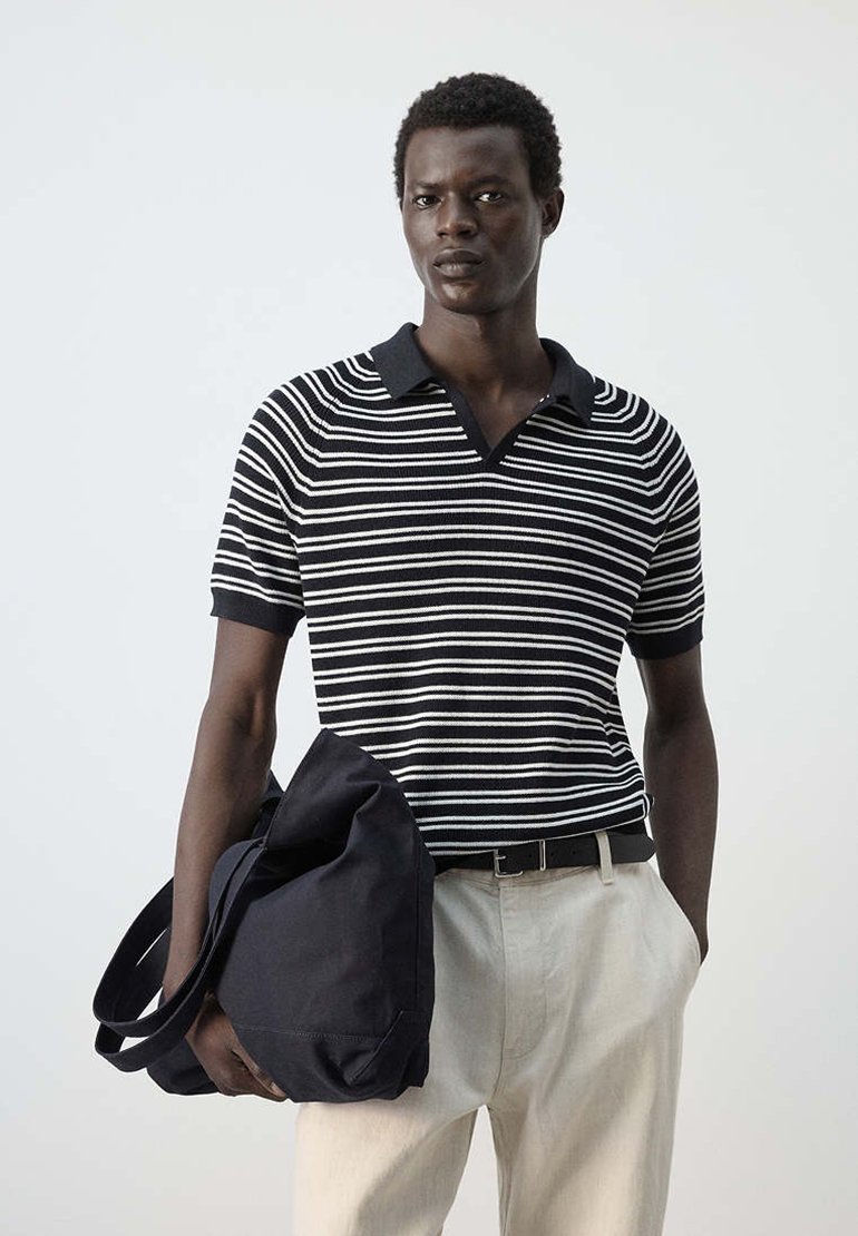 Homme portant un polo rayé noir et blanc et un pantalon clair, tenant un grand sac noir avec une main dans la poche.