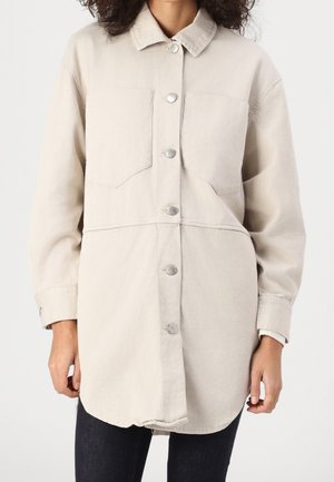 Vrouw in een beige knopen-overhemd met lange mouwen en grote voorzakken boven een donkere broek, staand tegen een effen achtergrond.