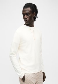 Mees dreadlockidega, seljas pikkade varrukatega kreemjas henley-särk ja beežide mustriliste pükstega, seisab käed taskutes, vaadates alla vasakule.