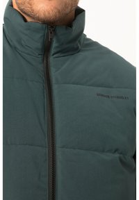 Dunkelgrüne Daunenjacke mit hohem Kragen und Frontreißverschluss, ausgestattet mit schwarzem Zubehör und Logo-Text im Brustbereich. Glatte Textur.