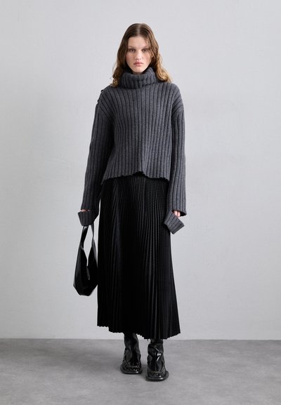 Suéter cinza canelado com gola alta, combinado com uma saia preta plissada e botas de cano alto pretas e brilhantes. Acessório de uma bolsa preta.