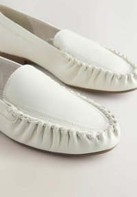 Baltos odinės slip-on bateliai suapvalintu priekiu, sutrauktomis kraštinėmis ir dekoratyviniu siuvinėjimu palei pado kraštą. Lygus paviršius, žemu kulniuku.