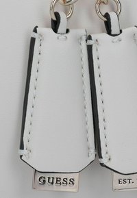 Witte leren labels met zwarte accenten, voorzien van zilverkleurige metalen hardware. De tekst omvat "GUESS" en "EST." met gestikte details.