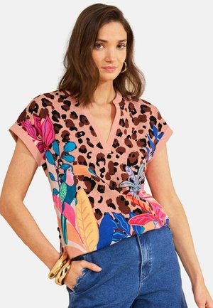 Femeie purtând o bluză cu mâneci scurte, imprimată cu model de leopard și frunze tropicale colorate și păsări, asortată cu blugi albaștri și brățări aurii.