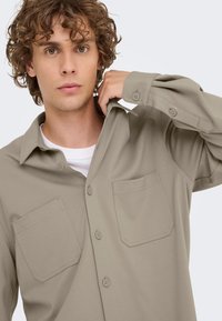 Lichtbruine blouse met knopen, een kraag, twee borstzakken en lange mouwen. Gemaakt van een gladde stof met subtiele stikseldetails.