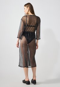 Copertura nera trasparente con un tessuto leggermente strutturato e scintillante; maniche tre quarti; lunghezza a metà polpaccio; vita elastica; indossata sopra il costume da bagno.