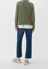 Olijfgroene gebreide trui over een wit shirt, gecombineerd met blauwe denim jeans en beige sneakers, gezien vanuit de achterkant.