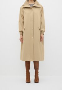 Abrigo largo beige con cuello alto, cierre de botones, mangas elásticas, bolsillos laterales y textura suave. Complementado por botas marrones hasta la rodilla.