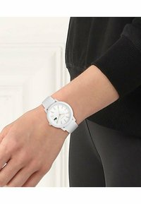 Montre blanche avec un bracelet en silicone texturé, cadran rond, accents dorés et verts, et marqueurs d'heure minimalistes sur le poignet d'une personne.