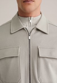 Helehall zipiga jakk soonikkoelise krae, kahe ees tasku ja tekstuurse kangaga. Omaduseks on hõbedane tõmblukk ja vormitud õlad.