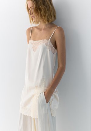 Femme aux cheveux blonds mi-longs portant un caraco blanc bordé de dentelle et un pantalon ample assorti, debout contre un mur clair.