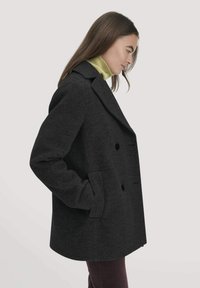 Doppelreihiger schwarzer Wollmantel mit Reverskragen und Seitentaschen, getragen über einem hellgrünen Rollkragenpullover. Glatte Textur, lässige Passform.