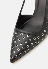 Escarpin noir à talon aigu, avec un bout pointu, présentant plusieurs œillets en métal argenté sur le dessus, et un design de talon ouvert et courbé. Texture en cuir lisse.