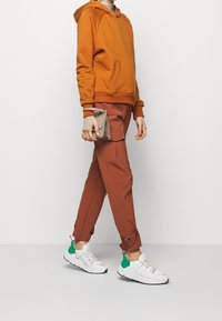 Sudadera naranja, capa inferior de punto beige, pantalones de color óxido con bolsillos laterales y zapatillas blancas con acentos verdes. Pequeño bolso beige en la mano.