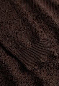Hnědá pletená látka s propracovanými krajkovými vzory a detailem žebrovaného lemu.