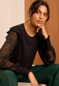 Top nero con maniche trasparenti ricamate che presentano motivi floreali, abbinato a pantaloni verdi e dettagli testurizzati.