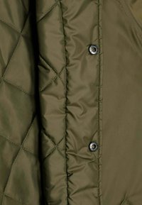 Tissu matelassé vert olive avec piqûres en losange et boutons-pression en métal sur une section de veste ou manteau rembourré.