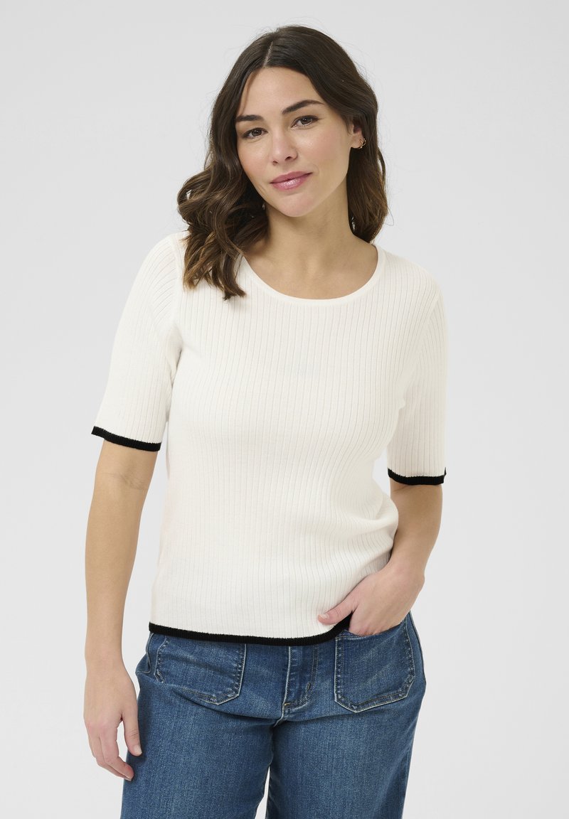Pulover crem cu nervuri, cu manșete scurte, având margini negre la manșete și la tiv; purtat cu blugi albaștri la talie înaltă. Design simplu și ajustat.