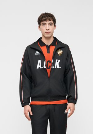 Jeune homme portant une veste de sport A.C.K. noire et orange avec un pantalon assorti, debout devant un fond clair uni.