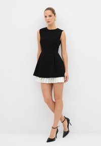 Robe noire sans manches avec un corsage ajusté et une jupe plissée blanche, associée à des escarpins noirs à bout pointu et à des boucles d'oreilles élégantes en or.