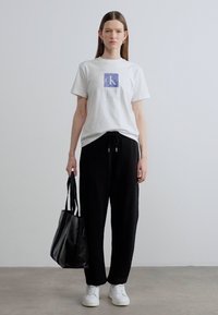 Λευκό βαμβακερό T-shirt με μπλε τετράγωνο λογότυπο, μαύρο παντελόνι με κορδόνι, λευκά αθλητικά παπούτσια και μαύρη τσάντα tote, παρουσιασμένα σε ουδέτερο φόντο.