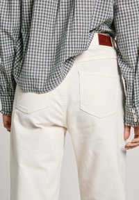 Pantalones de pana blancos con bolsillos traseros, que presentan una superficie ribeteada texturizada. Una camisa a cuadros en azul y blanco es parcialmente visible por encima.