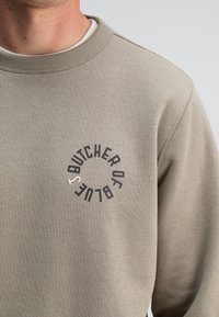 Lichtgrijze sweatshirt met een ronde hals. Bevat zwarte tekst "BUTCHER OF BLUTE" in een cirkelontwerp. Zachte textuur, geribbelde manchetten.
