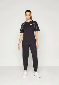 Μαύρη αθλητική στολή με κοντό μανίκι και λευκές λεπτομέρειες, συνοδευόμενη από ταιριαστά στενά παντελόνια. Φορεμένη με λευκά αθλητικά παπούτσια.