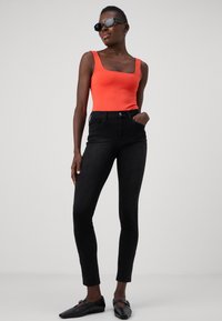 Oranje bodysuit met vierkante halslijn, gedragen met hoge zwarte skinny jeans. Het model draagt donkere zonnebrillen en zwarte platte schoenen. Neutrale achtergrond.
