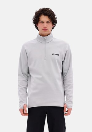 Joven con una sudadera deportiva gris claro con cremallera y logo "cep" y pantalones negros, posando frente a un fondo blanco.