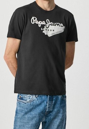 Mężczyzna ubrany w czarną koszulkę Pepe Jeans London z krótkim rękawem i niebieskie dżinsy, stojący z rękami za plecami na jednolitym tle.