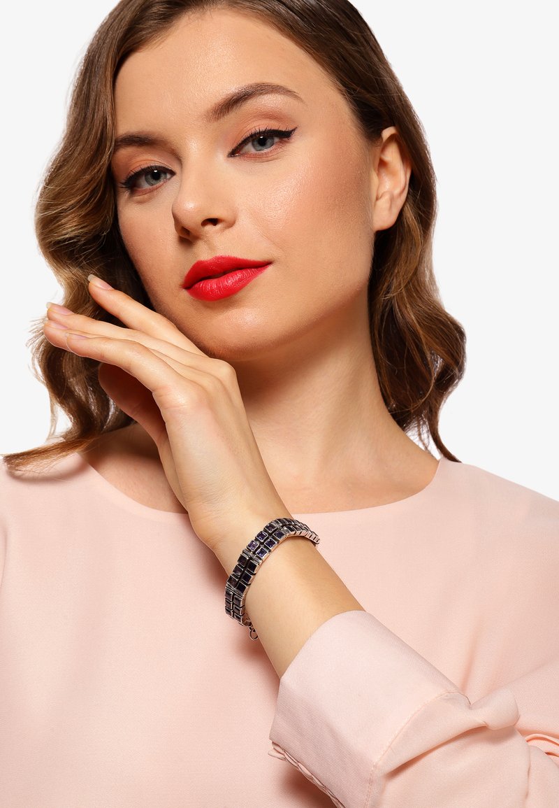 Silver armband med geometrisk design, med mörkblå accenter. Bäraren har ljus hy och vågigt brunt hår, klädd i en ljusrosa blus.