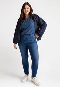 Chaqueta de mezclilla en navy con cierre de cremallera, combinada con una blusa azul y jeans ajustados azules con adornos. Zapatillas blancas completan el conjunto.